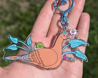 May include: A peach-colored bird with a green snail on its back, surrounded by blue leaves and glitter. The bird has a heart-shaped speech bubble with a pink heart inside. The bird is attached to a silver heart-shaped keychain.