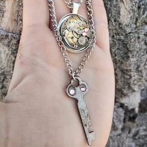 Puede incluir: Un collar de cadena de plata con dos colgantes. El colgante superior es un mecanismo de relojería redondo enmarcado en plata con diamantes de imitación rosas. El colgante inferior es una llave de plata con diamantes de imitación rosas.