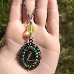 May include: A black chain necklace with a green glass bead and a black pendant with a green and iridescent crystal design. The pendant features a letter "Z" in the center.