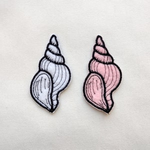 May include: Two embroidered fabric seashells, one white and one pink, with black outlines. The shells are in the shape of a conch shell.