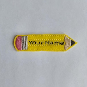 May include: A yellow felt pencil-shaped patch with the text "Your Name" embroidered in black. The pencil has a pink eraser, a gray ferrule, and a tan and black tip. The patch is designed for personalization.