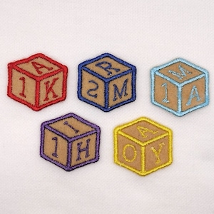 May include: Six embroidered felt blocks with the letters 'AK', '2M', '1A', '1H', 'OY', and '1K' in a variety of colors. The blocks are in the shape of a cube and are sewn onto a white background.