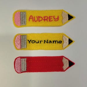 May include: Three felt pencil-shaped patches. The top patch is yellow with pink eraser, silver ferrule, and black tip, featuring the embroidered name Audrey in red. The middle patch is yellow with black text Your Name. The bottom patch is red.