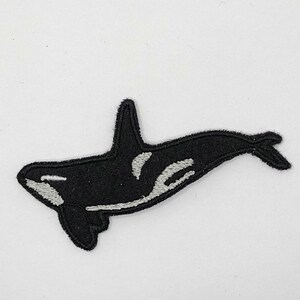 May include: Black and white orca patch. The patch features a detailed design of an orca whale with white accents on a black background. The patch is likely made of felt or a similar fabric and is designed to be sewn or ironed onto clothing or accessories.