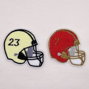 May include: Two embroidered iron-on patches of football helmets. One is yellow with the number 23 and the other is red with the number 7.