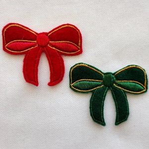 May include: Two embroidered bow appliques. One is red with gold trim, the other is green with gold trim. Both have a textured appearance and are set against a white background. These decorative bows could be used for crafts or holiday decorations.