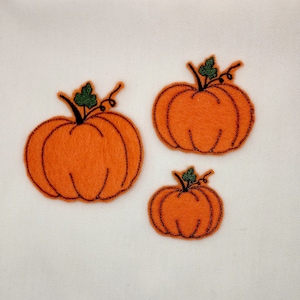 May include: Three orange felt pumpkin appliqués with dark brown stitched outlines and green leaf accents. The pumpkins vary in size and are arranged on a white background. These decorative patches are ideal for fall-themed crafts and decorations.