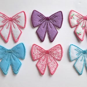 May include: Six embroidered fabric bow patches in various colors and patterns. The bows are pink, purple, blue, and white with floral patterns.