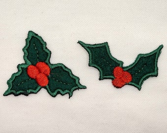 May include: Two embroidered holly leaf patches featuring dark green leaves with jagged edges and bright red berries. The left patch has three leaves and three berries, while the right patch has two leaves and three berries, all with black stitching.