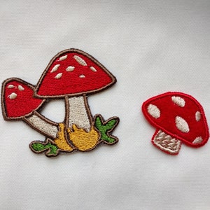 May include: Embroidered patches featuring red mushrooms with white spots. One patch shows two mushrooms clustered together, while the other depicts a single mushroom. The patches have brown, beige, yellow, and green accents.