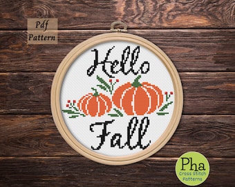 May include: Cross stitch pattern featuring the text "Hello Fall" with three orange pumpkins and green leaves. The pattern is displayed in a wooden embroidery hoop.