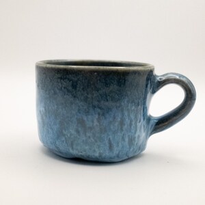 Peut inclure: Une tasse en céramique faite à la main avec un glaçage bleu texturé. La tasse a un corps arrondi et une anse incurvée. La couleur passe d'un bleu foncé au bord à un bleu plus clair vers la base. La tasse est conçue pour les boissons chaudes.