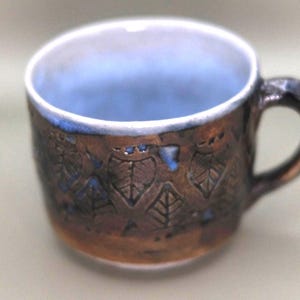 Peut inclure: Une tasse en céramique faite à la main avec un extérieur marron et un intérieur bleu. La tasse présente un motif de feuilles et une anse. Le bord est blanc avec un accent bleu. La tasse convient aux boissons chaudes ou froides.