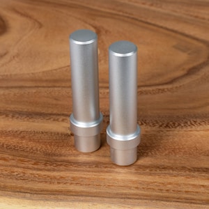 Könnte beinhalten: Zwei silberfarbene Metallzylinder mit flacher Oberseite und breiterem Sockel, die auf einer Holzoberfläche stehen. Die Objekte haben eine glatte, metallische Oberfläche und ein minimalistisches Design. Der Hintergrund ist eine warmtonige Holzmaserung. Ca. 5 cm hoch.