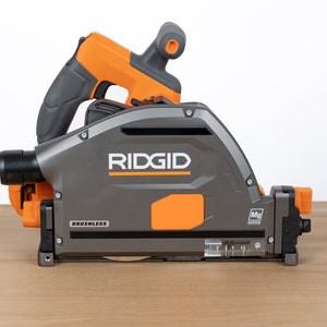 Puede incluir: Una sierra circular inal&aacute;mbrica Ridgid gris y naranja con motor sin escobillas. La sierra tiene un puerto de recolecci&oacute;n de polvo transparente y un indicador de profundidad transparente. La sierra est&aacute; sobre una superficie de madera.