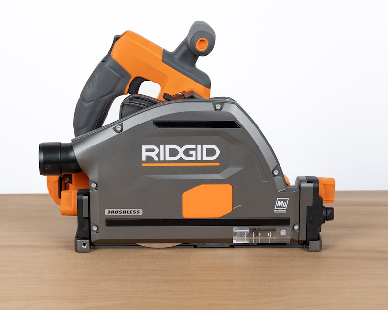 Puede incluir: Una sierra circular inal&aacute;mbrica Ridgid gris y naranja con motor sin escobillas. La sierra tiene un puerto de recolecci&oacute;n de polvo transparente y un indicador de profundidad transparente. La sierra est&aacute; sobre una superficie de madera.