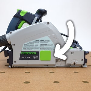 May include: A Festool TSV 60 KEB track saw with a green and black colour scheme. The saw is on a brown surface with a white background. The saw has a safety guard and a kickback stop.