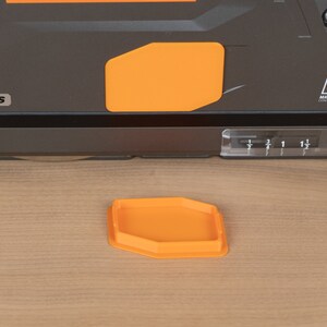 Puede incluir: Primer plano de una herramienta el&eacute;ctrica negra y naranja con un medidor de profundidad de pl&aacute;stico transparente. La herramienta tiene una etiqueta que dice "Mg Magnesium Construction Alloy". Una peque&ntilde;a bandeja hexagonal naranja est&aacute; sentada sobre una superficie de madera.