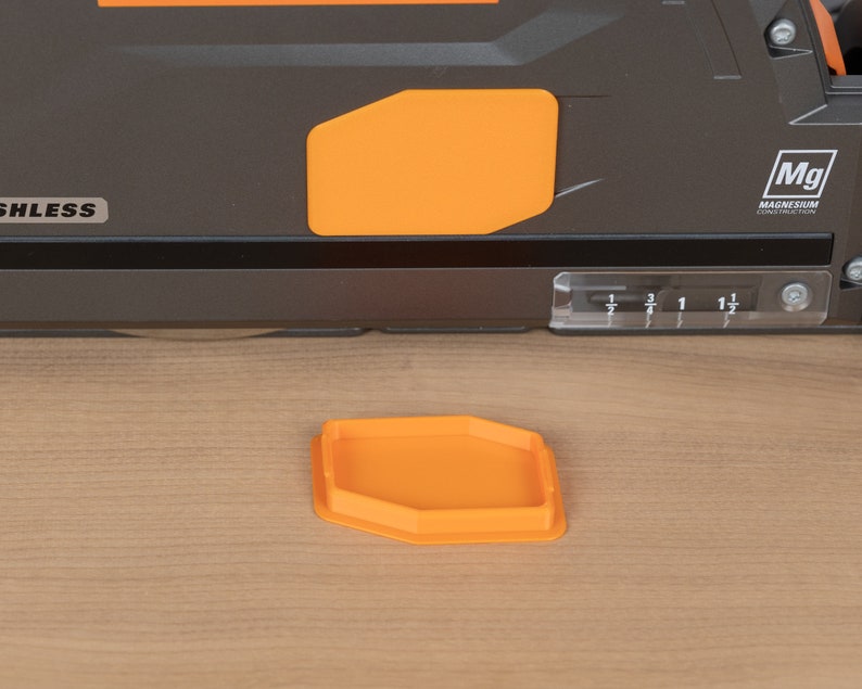 Puede incluir: Primer plano de una herramienta el&eacute;ctrica negra y naranja con un medidor de profundidad de pl&aacute;stico transparente. La herramienta tiene una etiqueta que dice "Mg Magnesium Construction Alloy". Una peque&ntilde;a bandeja hexagonal naranja est&aacute; sentada sobre una superficie de madera.