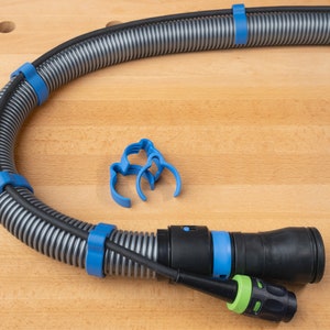 Può includere: Un tubo grigio avvolto con fascette per cavi blu. Il tubo ha un ugello nero con un connettore verde. Ci sono tre fascette per cavi blu aggiuntive su una superficie di legno.