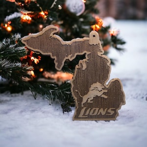 May include: A wooden ornament shaped like the state of Michigan with a lion logo and the word "LIONS" underneath. The ornament is sitting on a snowy surface.