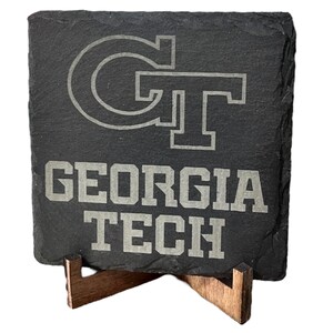 May include: A square, dark gray slate coaster with the Georgia Tech logo and the words "GEORGIA TECH" etched in white. The coaster is displayed on a small, wooden stand.