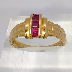 Può includere: Un anello d'oro con un design rettangolare con tre pietre preziose rosse. L'anello ha una fascia strutturata e uno stile classico ed elegante. Le pietre preziose sono disposte in fila.