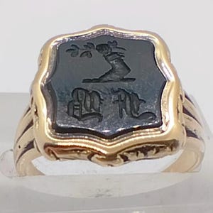 May include: A gold ring with a dark grey, shield-shaped centrepiece. The centrepiece features an engraved design of a person, a tree branch, and two stylized letters. The ring has a detailed, ornate design.
