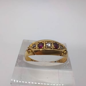 Może przedstawiać: Złoty pierścionek z rzędem czerwonych i białych kamieni szlachetnych. Pierścionek ma ozdobny wzór na obrączce.