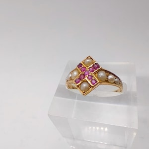 Könnte beinhalten: Ein goldener Ring mit einer rautenförmigen Fassung, die ein pinkfarbenes Rubinkreuz zeigt, das von weißen Perlen umgeben ist.