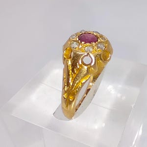 May include: A gold ring with a central, round, deep red gemstone encircled by smaller, clear stones. The ring's band and setting have an ornate floral design. The ring is presented on a clear, angled surface.