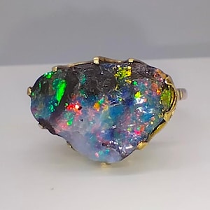 May include: A ring featuring a large, raw, irregular-shaped opal gemstone set in a gold-toned metal prong setting. The opal displays a dark base color with vibrant, multi-colored flashes of green, blue, red, and yellow throughout its surface.