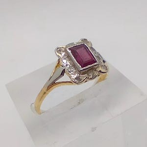 May include: A vintage gold ring featuring a rectangular, deep red gemstone at the center, surrounded by smaller, clear stones. The ring has a decorative, ornate setting and a two-tone gold and silver band. A classic piece of jewelry.