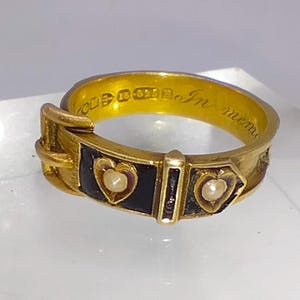 Könnte beinhalten: Ein goldener Ring mit einem schwarzen Emailleband und zwei kleinen Perlenherzen. Die Inschrift auf der Innenseite des Bandes lautet "In mem".