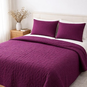 May include: A deep purple quilted bedspread and two matching pillowcases. The bedding is arranged on a bed with a light-coloured headboard. A wooden bedside table with a vase of dried flowers is visible.