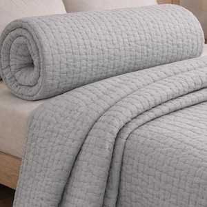 May include: A light grey quilt featuring a consistent square grid pattern throughout the fabric. The quilt is partially rolled up at the top, revealing the textured stitching and the soft, uniform colour of the textile material across the surface.