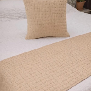 May include: A square beige throw pillow and a rectangular beige bed runner, both featuring a quilted grid pattern with visible stitching. The items are placed on a white quilted bedspread, showcasing a consistent light tan color and texture.