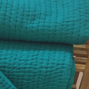 May include: A folded teal-colored quilt with a stitched, grid-like pattern. The fabric appears soft and textured, with visible seams creating a subtle, geometric design. The quilt is folded over itself, suggesting a cozy, comfortable item.