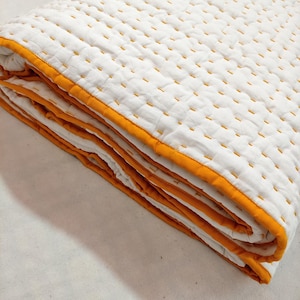 Op de afbeelding: Gevouwen witte quilt met gele stiksels en een gele rand. De quilt is gemaakt van een zacht, gewatteerd materiaal en is netjes opgevouwen. De stiksels zijn gelijkmatig verdeeld over het oppervlak, waardoor een textuurpatroon ontstaat.