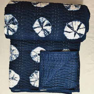 May include: A navy blue and white quilt with a tie-dye pattern. The quilt features large, circular white designs against a dark blue background. The stitching is visible in white thread, adding texture. The quilt is folded, showing its layered construction.