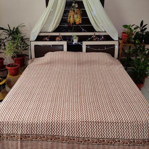 Puede incluir: Una cama cubierta con un edredón crema con un patrón de zigzag repetido en un tono rojo intenso. El edredón tiene un borde decorativo con un diseño floral. La cama está colocada contra un cabecero marrón oscuro.