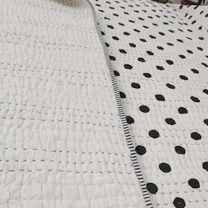 May include: White quilted blanket with black polka dots. The blanket has a textured surface and is bordered with a black and white striped trim. The design is simple and modern, suitable for a bedroom or living room.