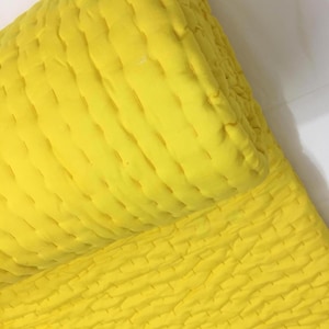 May include: A bright yellow quilted blanket featuring a textured surface with visible running stitch patterns throughout the fabric. The textile is folded over, displaying a soft, layered appearance with consistent stitching lines across the material.