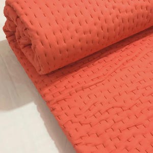 May include: A solid coral-colored textile featuring a repeating pattern of small, vertical running stitches across the entire surface. The fabric is folded into a thick, cylindrical roll, showcasing the textured, quilted appearance of the material.
