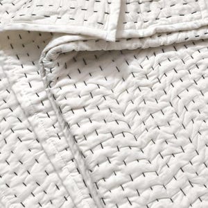 May include: A white quilted blanket featuring a repeating pattern of short, horizontal black stitched lines. The fabric has a textured, crinkled appearance with visible seams and a finished border edge, creating a consistent grid-like design.