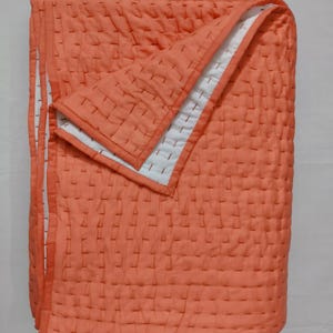 May include: A folded rectangular quilt featuring a vibrant coral orange top layer with visible white running stitch quilting patterns. The underside of the quilt is white, and the edges are finished with a matching coral orange fabric binding.
