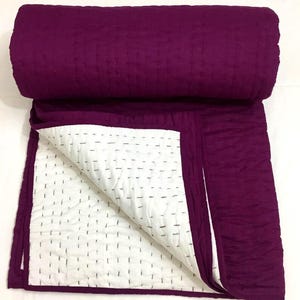 May include: A rolled-up, deep purple quilt with a white interior. The quilt is made of a textured fabric and features a stitched pattern. The white interior has a grid-like pattern of black stitching. The quilt is partially unrolled, revealing the white interior.