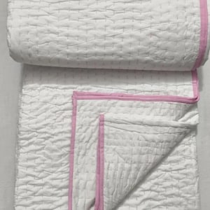 Pode incluir: Cobertor branco acolchoado com acabamento rosa, dobrado e enrolado. O cobertor tem uma superfície texturizada com um padrão quadriculado. A borda rosa contorna as bordas, adicionando cor.