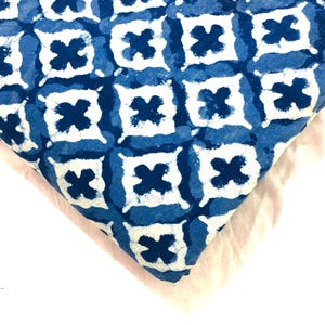 May include: A folded piece of fabric featuring a repeating pattern of white four-pointed cross shapes centred within blue squares. The design has a hand-dyed appearance with soft, irregular edges on the white motifs against a deep blue background.