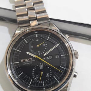 Puede incluir: Un reloj automático Seiko Chronograph de acero inoxidable con esfera negra. La esfera presenta marcas blancas y amarillas, tres subesferas y una pantalla de día/fecha. La correa del reloj es de acero inoxidable.